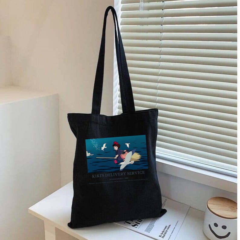 May include: Black tote bag featuring a colorful graphic of a witch flying on a broom over water, with the text "Kiki's Delivery Service". The bag has long black handles and is made of a sturdy fabric, perfect for carrying books or groceries.