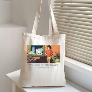 May include: Off-white canvas tote bag featuring a colorful anime scene from "Whisper of the Heart." The image shows two characters, a boy and a girl, with text that reads "Whisper of the Heart" and "Studio Ghibli 1995."