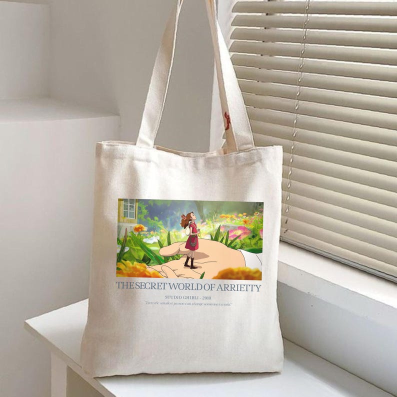 K&ouml;nnte beinhalten: Cremefarbene Tragetasche mit einer farbenfrohen Illustration aus "Arrietty &ndash; Die wundersame Welt der Borger". Das Bild zeigt ein M&auml;dchen, das auf einer riesigen Hand steht, umgeben von Blumen. Der Text "THE SECRET WORLD OF ARRIETTY" und "STUDIO GHIBLI - 2010" ist darunter gedruckt.