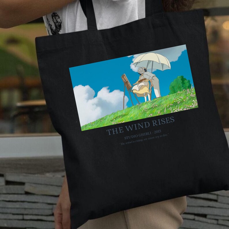 May include: Black tote bag featuring a colorful illustration from the animated film "The Wind Rises." The image shows two figures under an umbrella on a grassy hill with blue skies. Text on the bag reads "The Wind Rises" and "Studio Ghibli 2013."