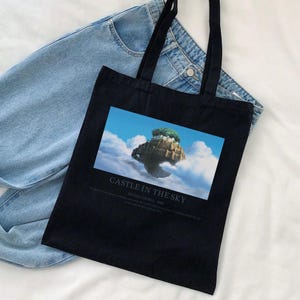 May include: Black tote bag featuring a colorful image of a floating island with the text "CASTLE IN THE SKY". The bag has long black handles and is made of a sturdy fabric. Light blue denim jeans are in the background.