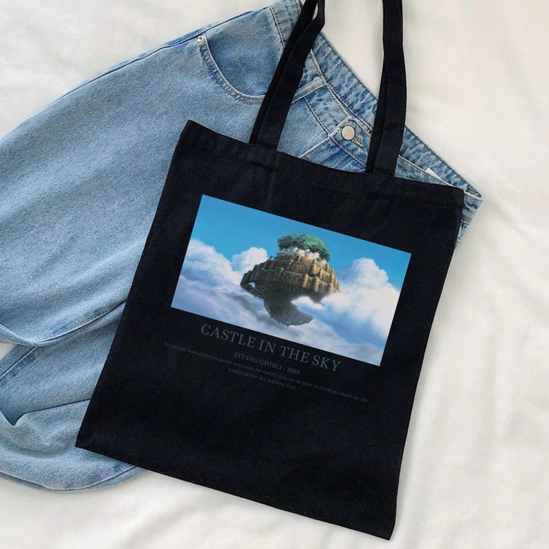 May include: Black tote bag featuring a colorful image of a floating island with the text "CASTLE IN THE SKY". The bag has long black handles and is made of a sturdy fabric. Light blue denim jeans are in the background.