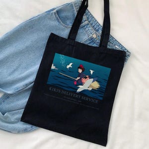 May include: Black tote bag featuring a colorful illustration of a young witch flying on a broom with a seagull. The text "Kiki's Delivery Service" is printed below the image. The bag has two black handles and is placed on top of a pair of blue jeans.