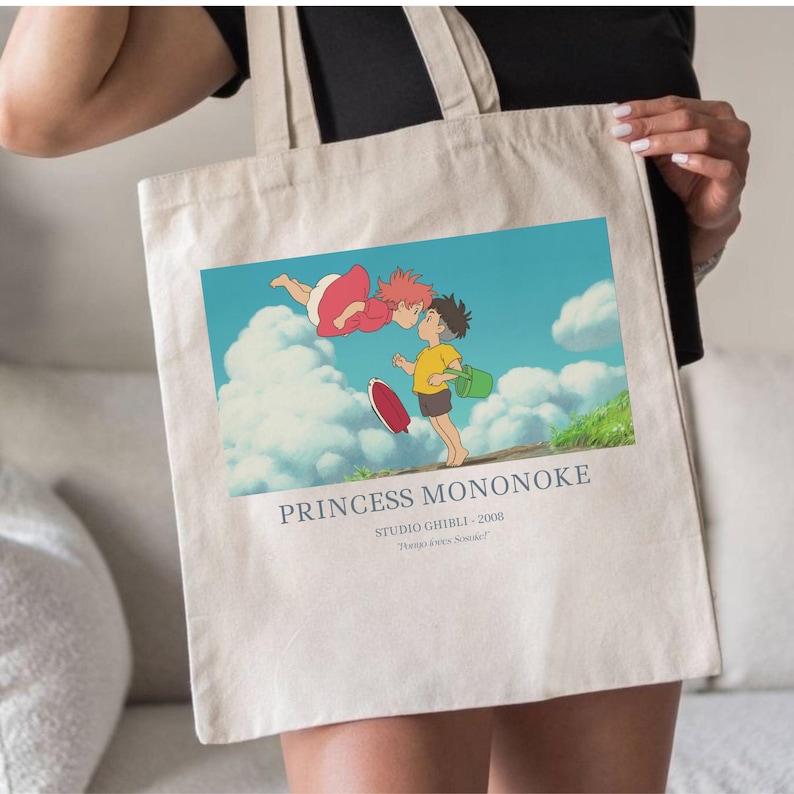 May include: Beige tote bag featuring a colorful illustration from the animated film "Princess Mononoke." The image shows two characters, one in a red dress and the other in a yellow shirt, with the text "PRINCESS MONONOKE" below the image.