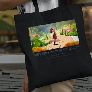 K&ouml;nnte beinhalten: Schwarze Tragetasche mit einer farbenfrohen Illustration aus "Arrietty &ndash; Die wundersame Welt der Borger". Das Bild zeigt ein M&auml;dchen, das auf einer riesigen Hand in einer lebendigen Au&szlig;enszene steht. Der Text auf der Tasche lautet "THE SECRET WORLD OF ARRIETTY" und "STUDIO GHIBLI - 2010."