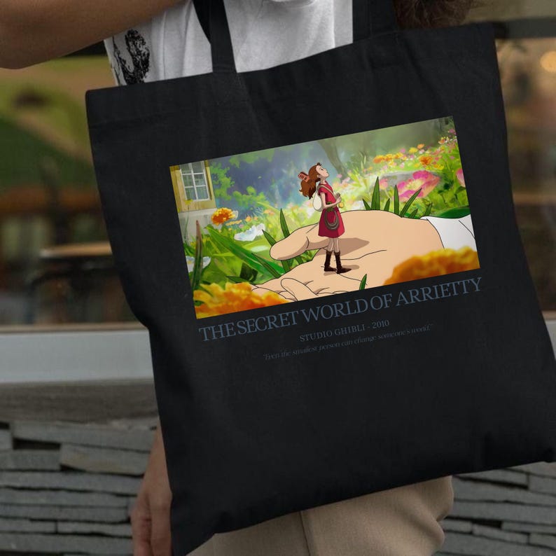 K&ouml;nnte beinhalten: Schwarze Tragetasche mit einer farbenfrohen Illustration aus "Arrietty &ndash; Die wundersame Welt der Borger". Das Bild zeigt ein M&auml;dchen, das auf einer riesigen Hand in einer lebendigen Au&szlig;enszene steht. Der Text auf der Tasche lautet "THE SECRET WORLD OF ARRIETTY" und "STUDIO GHIBLI - 2010."