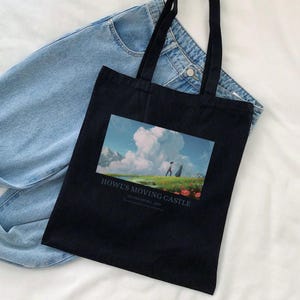 May include: Black tote bag featuring a colorful illustration from "Howl's Moving Castle." The image shows a landscape with a figure walking towards a castle under a cloudy sky. The bag has long black handles and is placed on top of a pair of blue jeans.