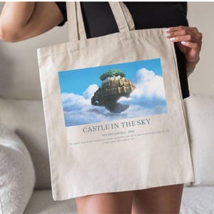 May include: Beige tote bag featuring a colorful image of a floating island with a castle and trees, set against a blue sky with clouds. The text "CASTLE IN THE SKY" and "STUDIO GHIBLI - 1986" are printed below the image.