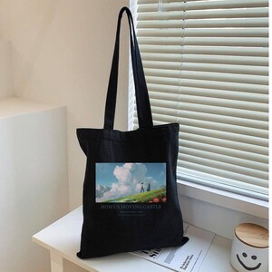 May include: Black tote bag featuring a colorful image from "Howl's Moving Castle." The image shows a person walking on a green hill with a blue sky and clouds. The words "Howl's Moving Castle" are printed below the image.