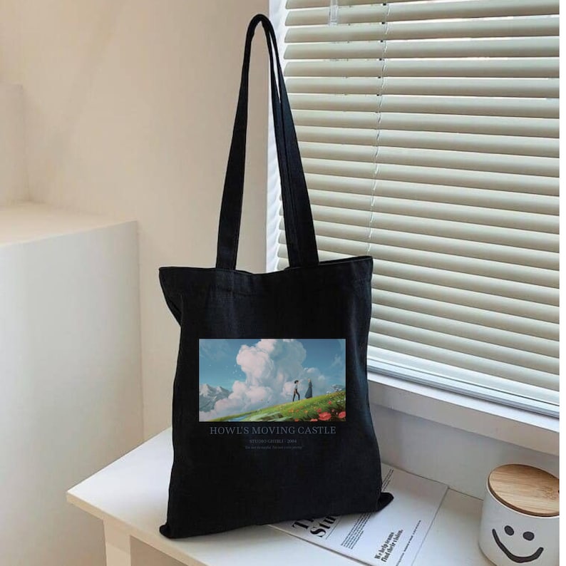 May include: Black tote bag featuring a colorful image from "Howl's Moving Castle." The image shows a person walking on a green hill with a blue sky and clouds. The words "Howl's Moving Castle" are printed below the image.
