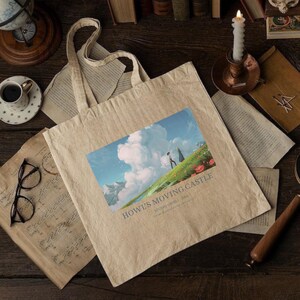 May include: Beige tote bag featuring a colorful image from "Howl's Moving Castle." The image shows a person walking on a grassy hill with a mountain backdrop and text that reads "Howl's Moving Castle Studio Ghibli 2004."