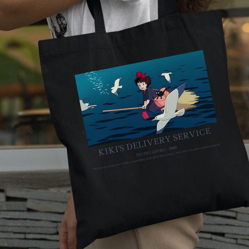 May include: Black tote bag featuring a colorful graphic from Kiki's Delivery Service. The image shows a young girl on a broom with a black cat, flying over water with seagulls. Text reads "Kiki's Delivery Service" and "Studio Ghibli - 1989."