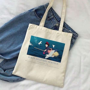May include: Off-white canvas tote bag featuring a colorful illustration from "Kiki's Delivery Service." The image shows a young witch flying on a broom over the ocean, with the text "Kiki's Delivery Service" below. The bag has two long handles and is placed on top of blue jeans.