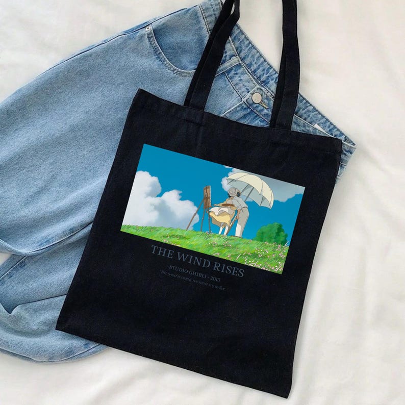 May include: Black tote bag featuring a colorful illustration from the Studio Ghibli film "The Wind Rises." The image shows a person under a parasol on a grassy hill. The text "THE WIND RISES" is printed below the image.
