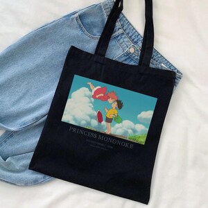 May include: Black tote bag featuring a colorful image from the animated film "Princess Mononoke." The image shows two characters in a sky scene. The text "PRINCESS MONONOKE" is printed below the image. The bag has black handles and is placed on top of a pair of blue jeans.