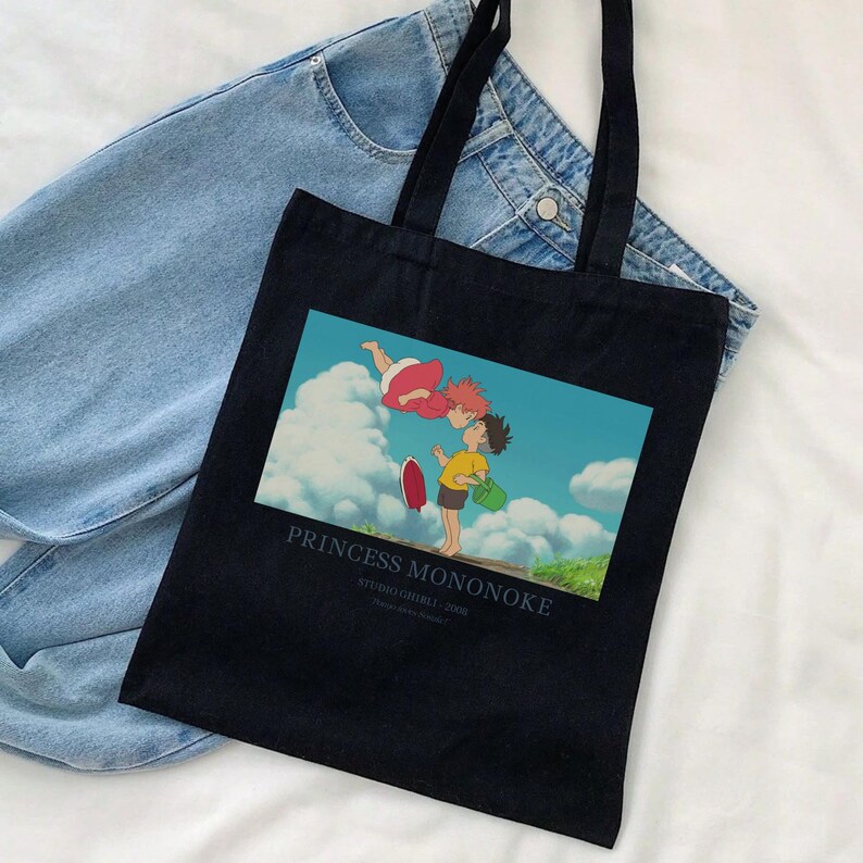 May include: Black tote bag featuring a colorful image from the animated film "Princess Mononoke." The image shows two characters in a sky scene. The text "PRINCESS MONONOKE" is printed below the image. The bag has black handles and is placed on top of a pair of blue jeans.