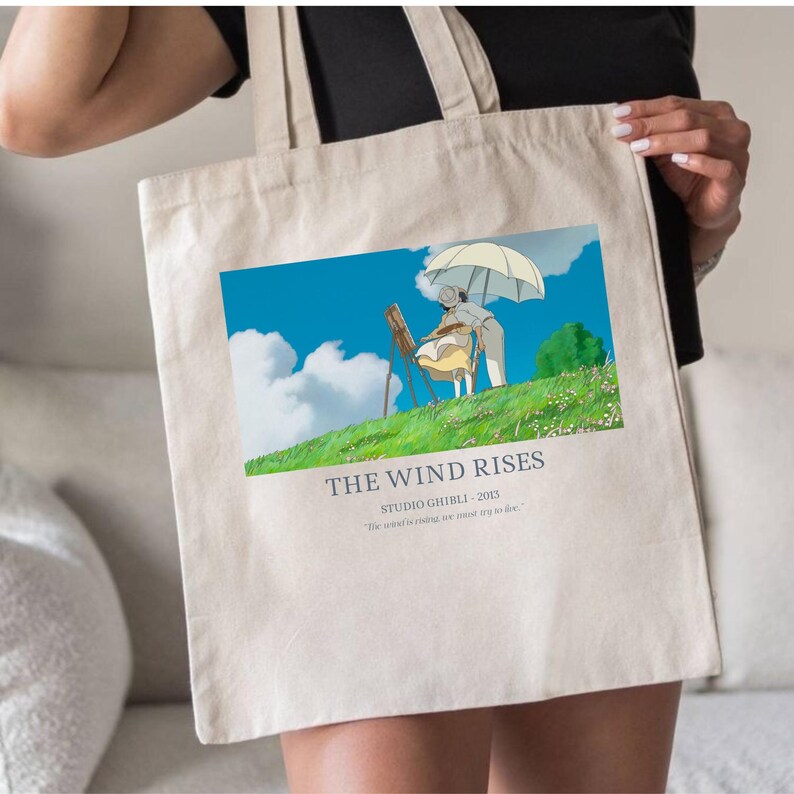 May include: Beige tote bag featuring a colorful illustration from "The Wind Rises" with a blue sky, clouds, and characters. The text "THE WIND RISES" and "STUDIO GHIBLI - 2013" are printed below the image. The bag is held by a person.