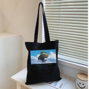 May include: Black tote bag with long handles. The bag features a rectangular image of a floating island in the sky, with the text "CASTLE IN THE SKY" below the image. The bag is sitting on a white surface.