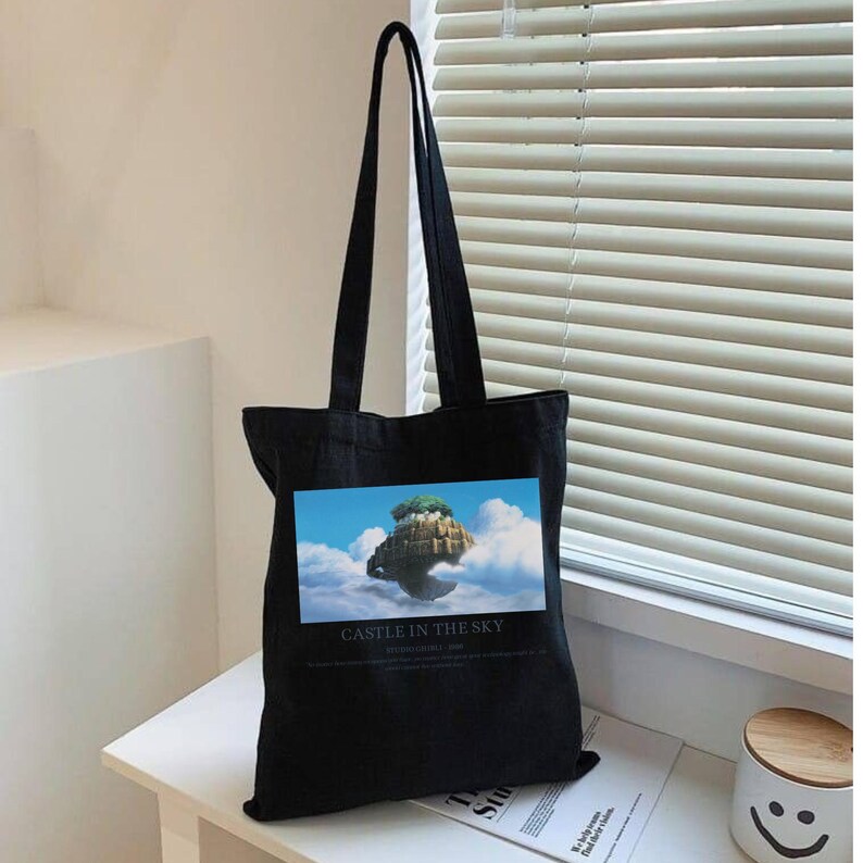 May include: Black tote bag with long handles. The bag features a rectangular image of a floating island in the sky, with the text "CASTLE IN THE SKY" below the image. The bag is sitting on a white surface.