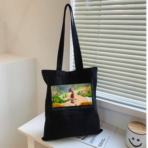 K&ouml;nnte beinhalten: Schwarze Tragetasche mit langen Henkeln. Die Tasche zeigt eine farbenfrohe Grafik eines M&auml;dchens, das auf einer Hand in einer Gartenszene steht. Der Text "THE SECRET WORLD OF ARRIETTY" ist darunter gedruckt.