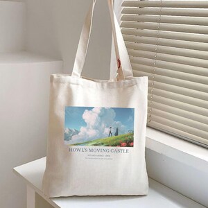 May include: A cream-colored tote bag featuring a colorful graphic from "Howl's Moving Castle." The image shows two figures on a grassy hill with a cloudy sky. Text on the bag reads "Howl's Moving Castle" and "Studio Ghibli - 2004."