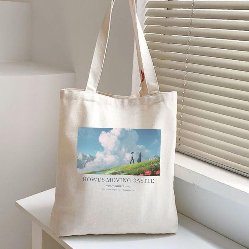 May include: A cream-colored tote bag featuring a colorful graphic from "Howl's Moving Castle." The image shows two figures on a grassy hill with a cloudy sky. Text on the bag reads "Howl's Moving Castle" and "Studio Ghibli - 2004."