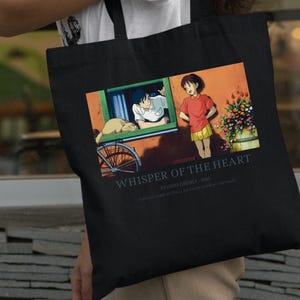 May include: Black tote bag featuring a colorful illustration from the animated film "Whisper of the Heart." The image shows two characters, a cat, and a bicycle. Text on the bag reads "Whisper of the Heart" and "Studio Ghibli - 1995."