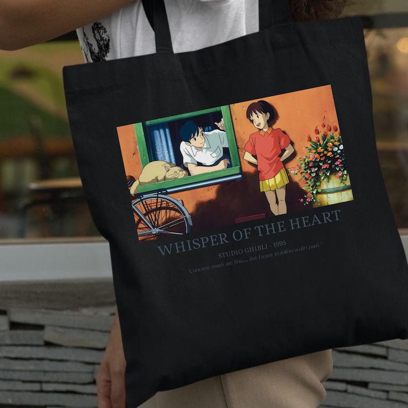May include: Black tote bag featuring a colorful illustration from the animated film "Whisper of the Heart." The image shows two characters, a cat, and a bicycle. Text on the bag reads "Whisper of the Heart" and "Studio Ghibli - 1995."