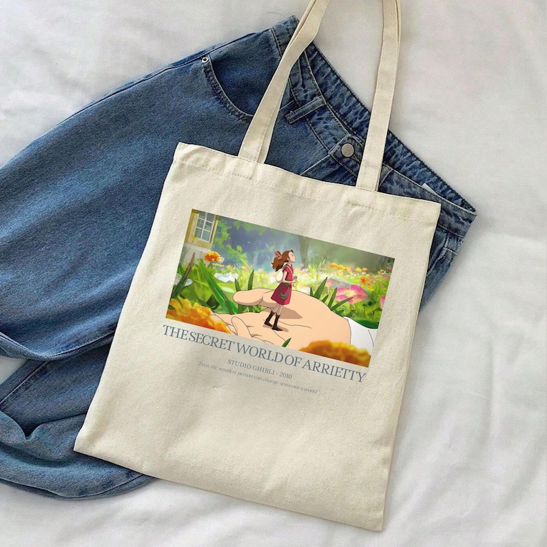 K&ouml;nnte beinhalten: Cremefarbene Tragetasche mit einer farbenfrohen Illustration aus "Arrietty &ndash; Die wundersame Welt der Borger". Das Bild zeigt ein M&auml;dchen, das auf einer Hand in einem Garten steht. Die Tasche hat lange Henkel und liegt auf einer Jeans.