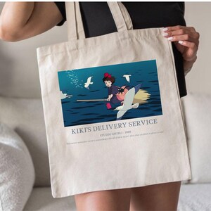 May include: Beige tote bag featuring a colorful graphic from Kiki's Delivery Service. The image shows a young witch flying on a broom over water, with seagulls. Text on the bag reads "Kiki's Delivery Service" and "Studio Ghibli - 1989".