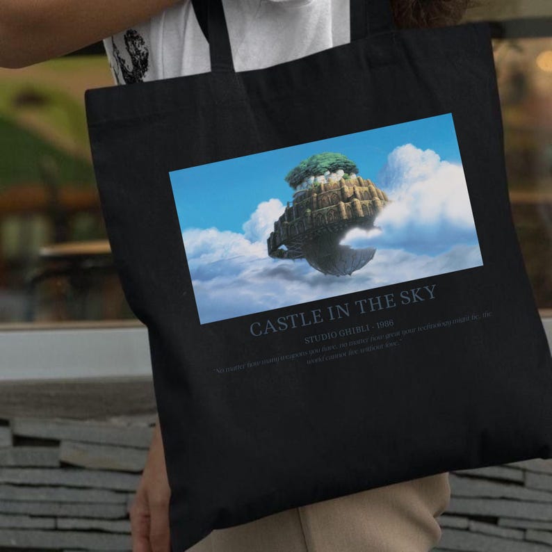 May include: Black tote bag featuring a colorful image of a floating island in the sky, with the text "CASTLE IN THE SKY" and "STUDIO GHIBLI - 1986". The bag is made of a durable fabric.