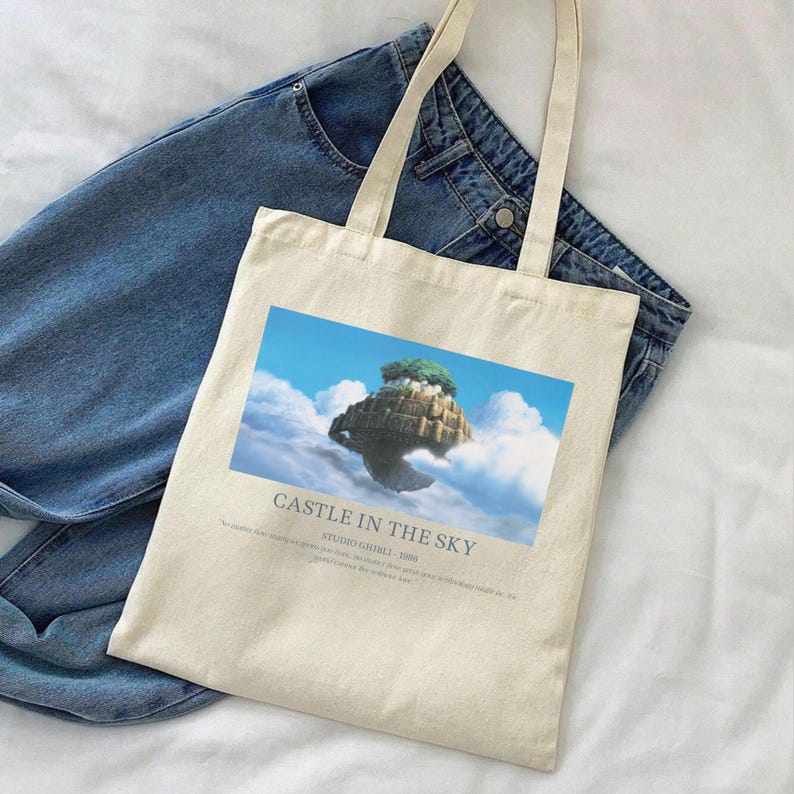 May include: Cream-colored tote bag featuring a graphic of a floating island with a castle and trees against a blue sky. The text "CASTLE IN THE SKY" is printed below the image. The bag has two long handles and is placed on top of blue jeans.