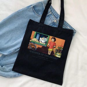 May include: Black tote bag featuring a colorful anime scene from "Whisper of the Heart." The image shows two characters, a boy and a girl, with the text "Whisper of the Heart" and "Studio Ghibli - 1995" printed below the image. The bag has black handles.