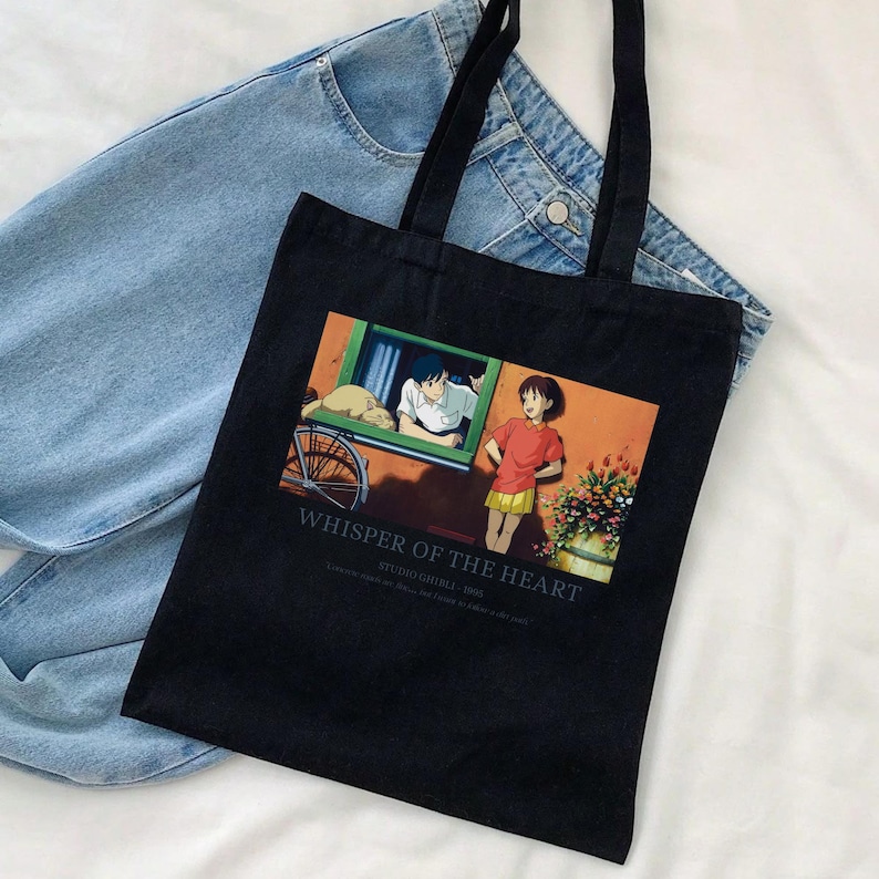 May include: Black tote bag featuring a colorful anime scene from "Whisper of the Heart." The image shows two characters, a boy and a girl, with the text "Whisper of the Heart" and "Studio Ghibli - 1995" printed below the image. The bag has black handles.