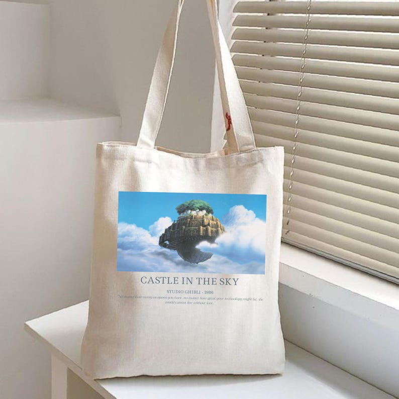 May include: Off-white canvas tote bag featuring a colorful image of a floating castle in the sky, surrounded by clouds. The text "CASTLE IN THE SKY STUDIO GHIBLI 1986" is printed below the image. The bag has two handles.