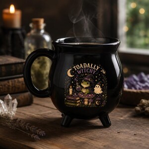 May include: A black ceramic cauldron-shaped mug with three small feet and a handle. The front features a colorful graphic of a frog wearing a witch hat, a ghost, a candle, and books, with the text "TOADALLY WITCHY" printed in a curved arch.