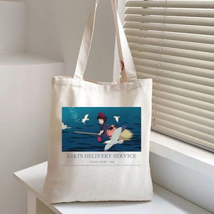 May include: A cream-colored tote bag featuring a colorful illustration from "Kiki's Delivery Service." The image shows a young girl on a broomstick with a black cat, flying over water with seagulls. Text at the bottom reads "Kiki's Delivery Service."