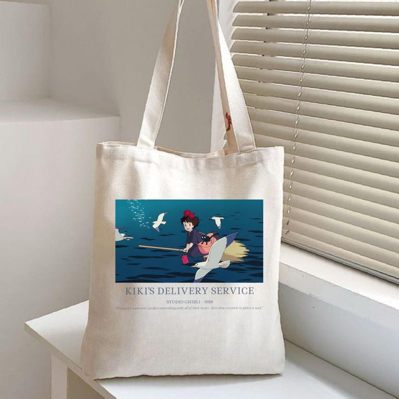 May include: A cream-colored tote bag featuring a colorful illustration from "Kiki's Delivery Service." The image shows a young girl on a broomstick with a black cat, flying over water with seagulls. Text at the bottom reads "Kiki's Delivery Service."