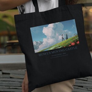 May include: Black tote bag featuring a colorful graphic from the animated film "Howl's Moving Castle." The image shows two figures on a grassy hill with a mountain backdrop and the text "Howl's Moving Castle" below.
