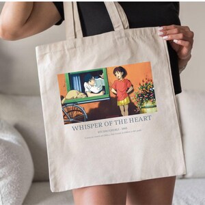 May include: Beige tote bag featuring a colorful graphic from the animated film "Whisper of the Heart." The image shows two characters, a bicycle, and the text "Whisper of the Heart - 1995." The bag is held by a person.