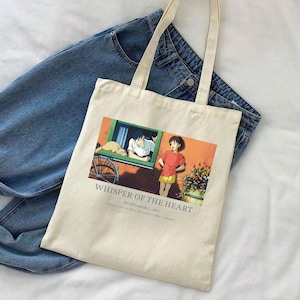 May include: Cream-colored tote bag featuring a colorful anime scene with the text "Whisper of the Heart." The bag has long handles and is placed on top of a pair of blue jeans. The graphic depicts two characters and a cat.