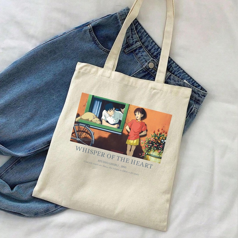 May include: Cream-colored tote bag featuring a colorful anime scene with the text "Whisper of the Heart." The bag has long handles and is placed on top of a pair of blue jeans. The graphic depicts two characters and a cat.