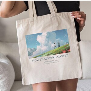 May include: Beige tote bag featuring a colorful illustration from the animated film "Howl's Moving Castle." The image depicts two figures on a grassy hill with a cloudy sky and text that reads "Howl's Moving Castle Studio Ghibli - 2004."