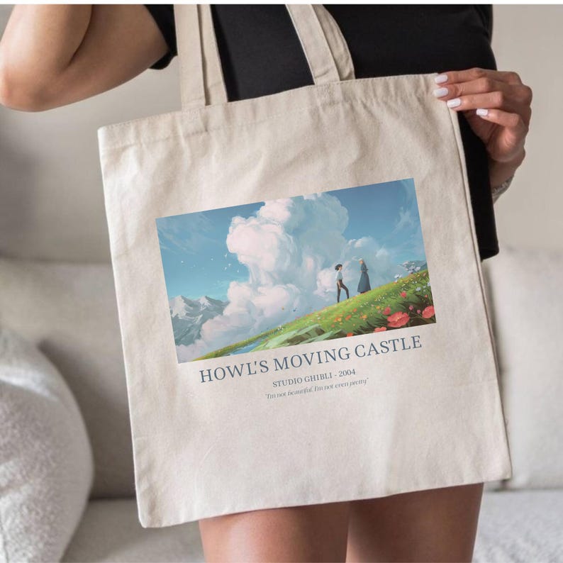 May include: Beige tote bag featuring a colorful illustration from the animated film "Howl's Moving Castle." The image depicts two figures on a grassy hill with a cloudy sky and text that reads "Howl's Moving Castle Studio Ghibli - 2004."