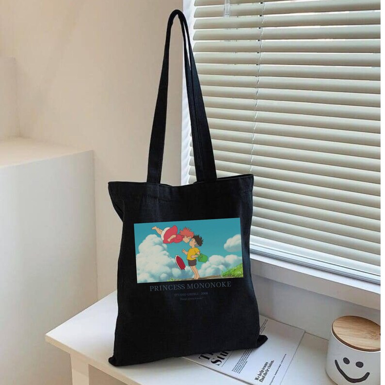 May include: Black tote bag featuring a colorful graphic from the animated film "Princess Mononoke." The image shows two characters in a sky scene. The bag has long handles and is made of a sturdy material, perfect for carrying books or groceries.