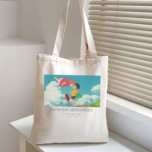 May include: Off-white canvas tote bag featuring a colorful illustration from the animated film "Princess Mononoke." The image shows two characters in a blue sky with clouds. The text "PRINCESS MONONOKE" is printed below the image.