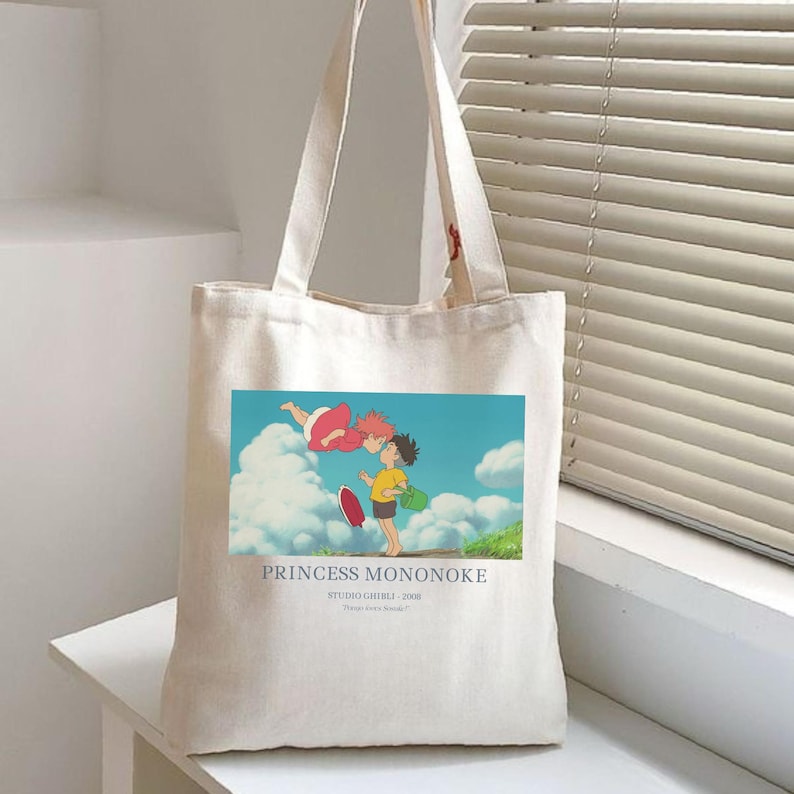 May include: Off-white canvas tote bag featuring a colorful illustration from the animated film "Princess Mononoke." The image shows two characters in a blue sky with clouds. The text "PRINCESS MONONOKE" is printed below the image.