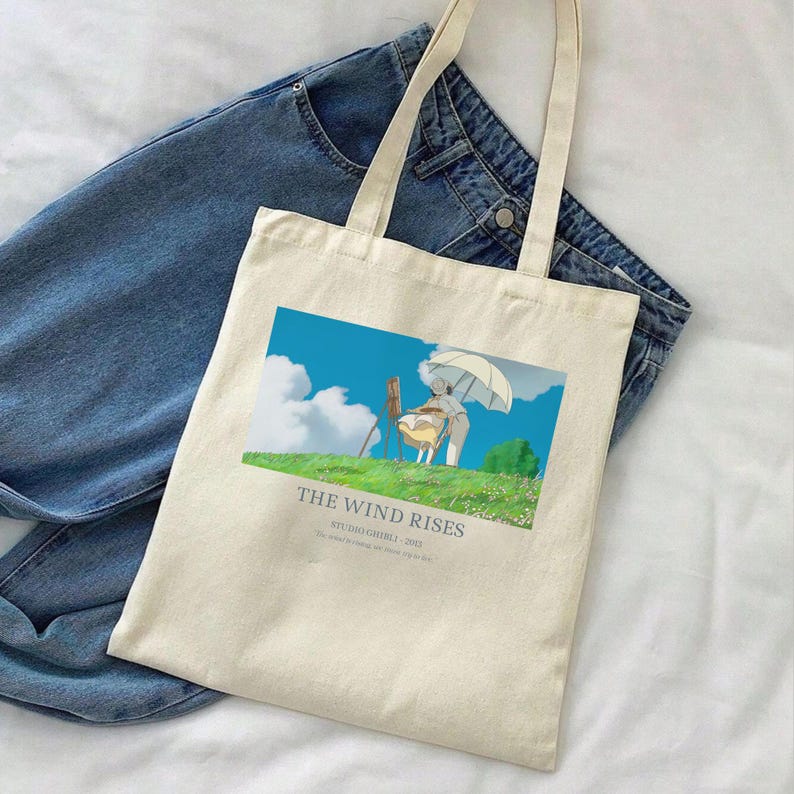 May include: Cream-colored tote bag featuring a colorful illustration from "The Wind Rises" with text. The bag has long handles and is placed on top of a pair of blue jeans. The illustration shows a person sitting on a chair under an umbrella.