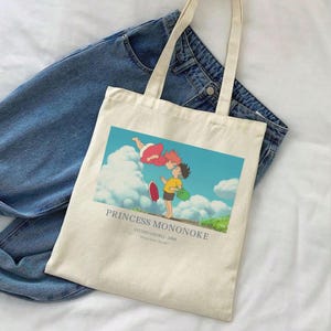 May include: Cream-colored tote bag featuring a colorful graphic from "Princess Mononoke." The image shows two characters with a blue sky and clouds background. The bag has long handles and is placed on top of a pair of blue jeans.