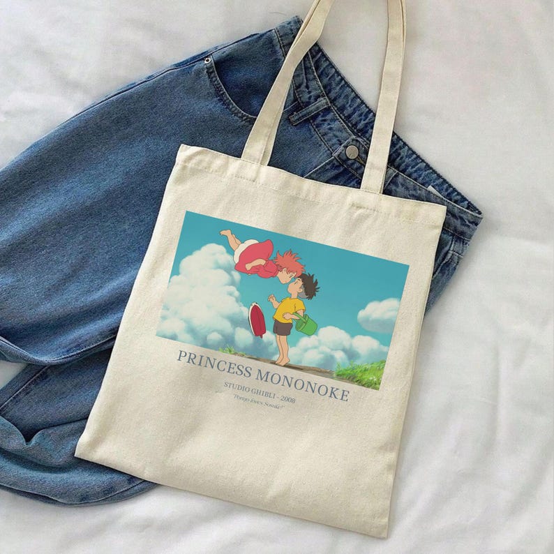 May include: Cream-colored tote bag featuring a colorful graphic from "Princess Mononoke." The image shows two characters with a blue sky and clouds background. The bag has long handles and is placed on top of a pair of blue jeans.